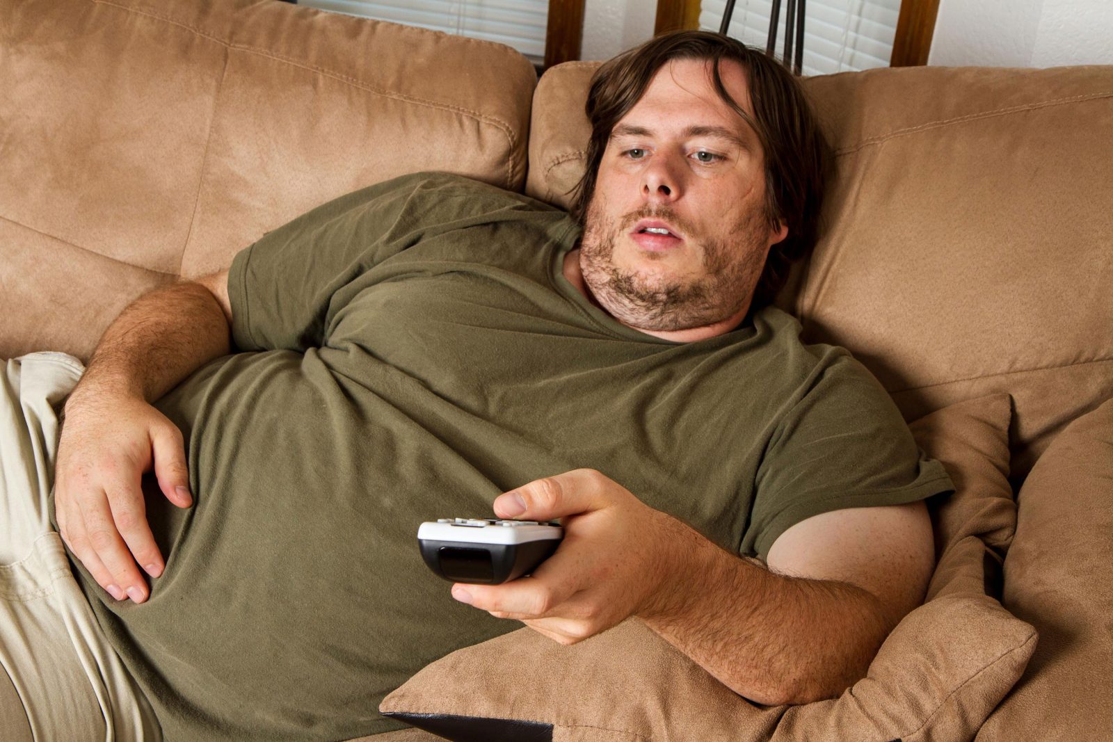 Толстый мужчина лежит на диване и переключает пульт от телевизора