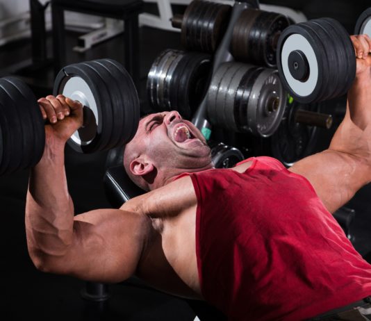 Опытный атлет в тренажерном зале выполняет упражнение: жим гантелей лежа на наклонной скамье