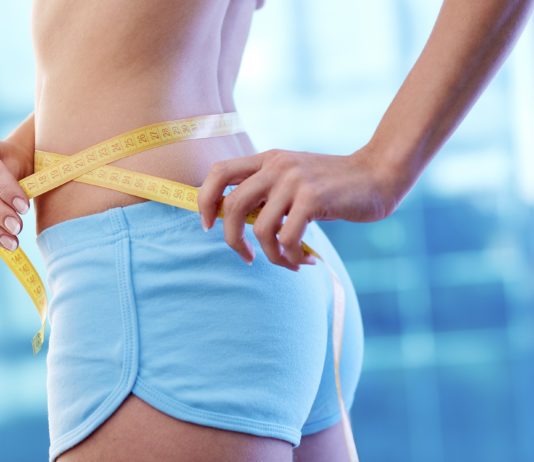 Как избавиться от лишнего веса? Стройная девушка в голубых шортиках измеряет талию сантиметровой лентой
