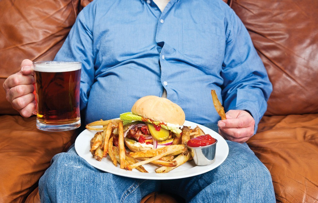 Толстый мужик пьет пиво на диване и ест вредные продукты