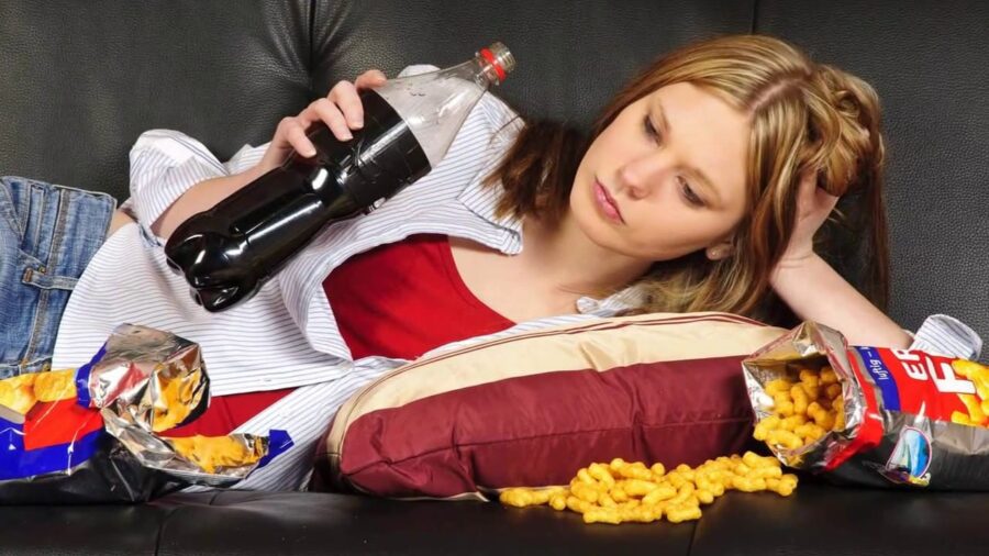 Девушка лежит на диване пьет газировку заедая фастфудом